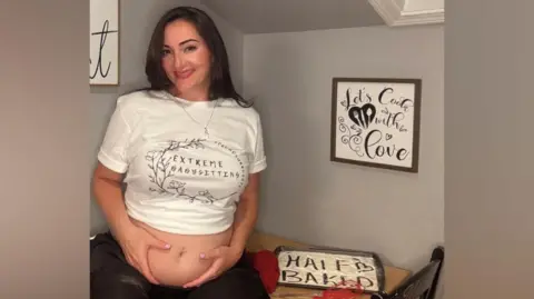 Krista Scelza is wearing a white cropped top, which reads "Extreme babysitting", to show her pregnant belly. To left is a sign on the wall which reads "Let' cook with love" and a baking tray with flour with the words "Half-baked". 