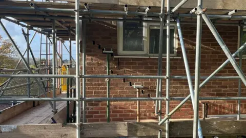 Scaffolding around a house. Cracks in the brickwork running down the side of the wall are visible