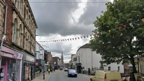Google Fisherton Street with shops, cars, pedestrians, a tree in the foreground on the right and a church spire in the background