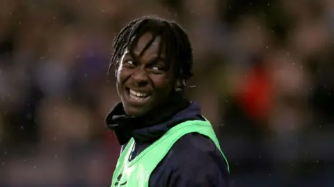 Quevin Castro is smiling as he looks over his shoulder. He has short dreadlocks and is wearing a tracksuit top and green bib.
