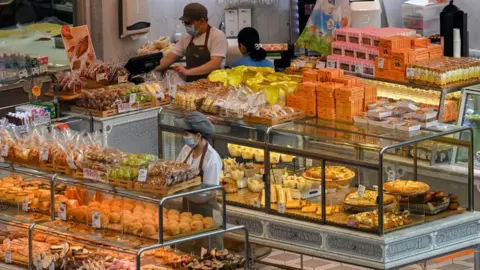 Getty Images A wide shot of a Singapore bakery filled with breads and doughnuts. A woman with a face mask and an apron is arranging the bread. Standing nearby her is another man, also in an apron, helping a customer at a cashier.