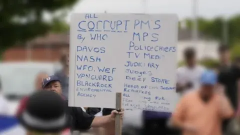 PA Media A man, wearing a blue England football hat, holds a white placard with blue writing. The writing says government ministers and MPs are corrupt, as are police chiefs, journalists, judges and the deep state, among a lot of other agencies.