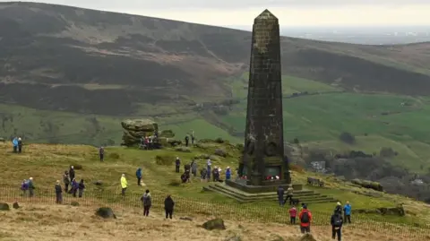 Scores of people gather while socially distancing during a memorial event in 2020 around the Saddleworth memorial. It is a tall slender obelisk pillar on a hill, surrounded by other hills in the Pennine countryside.