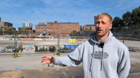 A man in a grey hoodie with his right arm outstretched stands in front of the demolished skatepark, which is fenced off.