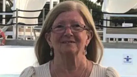 University Hospitals Birmingham NHS Foundation Trust A woman with long brown hair and glasses is looking at the camera. She is wearing a light top and a necklace.