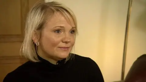 Elizabeth Hudson, a woman in her forties, with blonde hair, circular silver earrings, and a black turtle neck. It is a close-up shot of her head and shoulders, seemingly in her home or a living room.