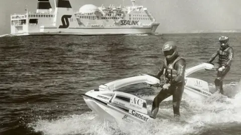 Lee Robinson A black-and-white photograph showing two men riding early-model stand-up jet skis through open water, with a large Sealink passenger ferry travelling in the background.