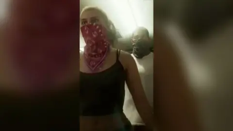 Still of GTA, a woman waring a red scarf over her mouth and a man behind her wearing a black scarf over his
