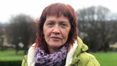 Paula Martin is an older woman with shoulder-length auburn hair and she is looking directly into the camera with a neutral expression on her face. She is stood in the middle of a park but the background is blurred. She is wearing a purple patterned scarf and a lime green puffer coat.