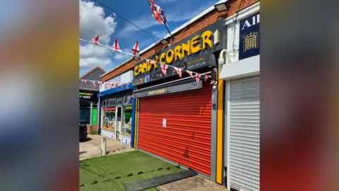 A shop called Candy Corner - with a red security shutter. Artificial grass matting is on the ground outside. There are St George's flags hung up either side of the shop front. 