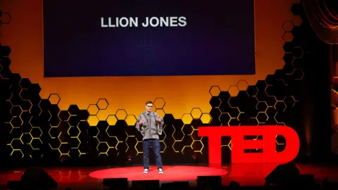 TED AI Llion Jones stands on a circular red carpet at a TED event, speaking to an audience. Behind him is a large screen displaying the name “LLION JONES” in bold white letters on a dark background. The stage backdrop features a honeycomb-like pattern in black and yellow tones. To the right of the speaker, large red letters spell out “TED.” 