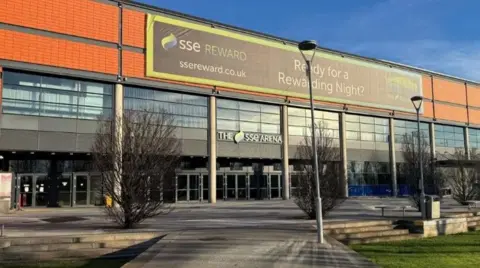 The outside of Belfast SSE Arena. A number of trees and lamp posts are visible outside the 11,000 capacity venue. A sign reading: 'SSE Reward' is visible on the top of the building.