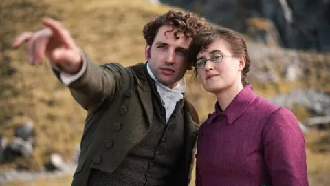 Laurie Davidson and Ella Brucolleri in a still from The Other Bennet sister. Laurie is a man with brunette curly hair, a dark green jacket, a brown waistcoat and a white cravat. he is pointing to the left. Ella is wearing a cropped pink jacket and a blue tartan skirt. 