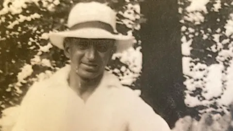 Winstones A black and white picture of a man standing in front of a tree. The man is wearing a white shirt and white wide-brimmed hat, and is smiling at the camera.