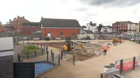 A triangular shaped area, which was once a shallow paddling pool, is now an area of rubble. A digger and small tipper truck are on site and a man in an orange hi-vis jacket and hard hat is in the foreground. There are railings around the paddling pool area.