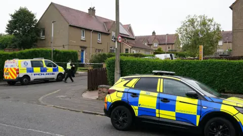 PA Media Police at crime scene in Edinburgh. Two police cars are packed at the end of a street of small, brown-harled houses. There is police tape over the entrance to the street.