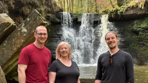 Johnny Reed/BBC A group of three people, two men and a woman, stand in a bright forest setting, with a cascading waterfall visible behind them