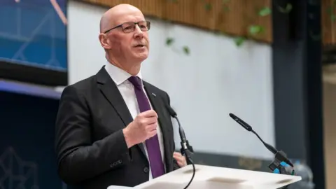 Getty Images John Swinney berbicara di podium putih. Dia mengenakan setelan gelap, kemeja putih, dan dasi ungu dengan tangan kanannya dipegang di depan dadanya. 