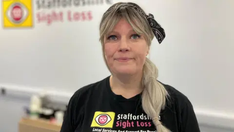 A woman with long blonde hair, tied back with a black bandana and in a pony tail which is draped over her left shoulder, smiles as she stands in a room, in front of a white wall which says Staffordshire Sight Loss. She is wearing a black shirt which says the charity's name and has the logo next to it.