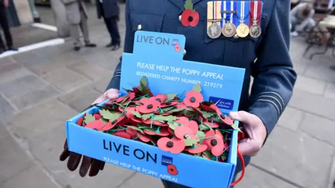 A veteran carrying a poppy appeal box with poppies inside it. The man is dressed in a blue uniform with a poppy and a number of medals. 
