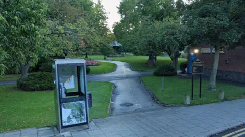 Google Selby park is pictured from the outside, with a phone box present in the left foreground. There's a path leading into the park in the middle, with a small black sign to the right. Trees are on either side of the path, and there are bushes and flowers. A bandstand and a bench can be seen in the distance.