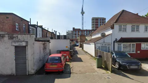 A narrow residential alleyway in an urban neighbourhood, viewed from the street. In the foreground, a red hatchback car is parked beside a small white garage with a closed metal roller door. A bin is on the right side of the alley near a wooden fence and a white semi-detached house with a tiled roof. A dark-coloured car is parked in a driveway to the right. Further along the alley, additional bins and low-rise buildings are visible, with a taller modern apartment block and a construction crane rising in the background against a clear blue sky. The scene is lit by bright sunlight, casting sharp shadows along the ground and walls.