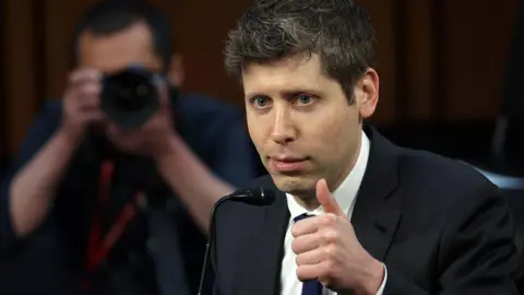 Sam Altman wearing a dark suit and tie, giving a thumbs up