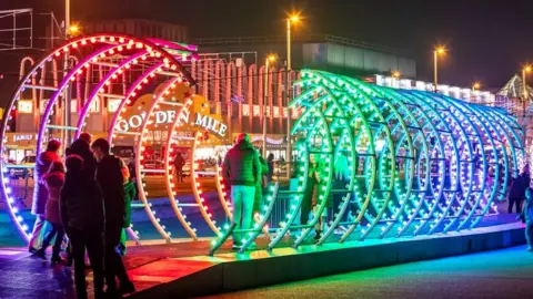 VisitBlackpool A large light installation, in rainbow shades, resembling a giant Slinky. People are standing at one end and a man is walking through it.