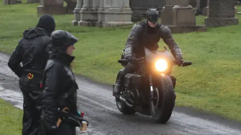 Filming on a scene in The Batman - a man dressed in a Batman costume is riding a motorbike in a graveyard, watched by crew members