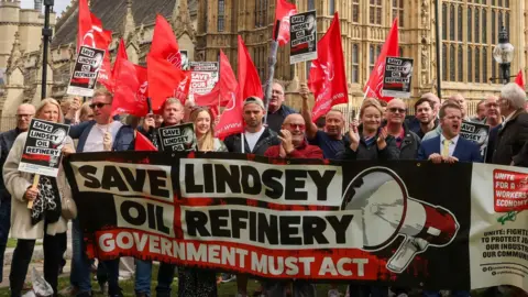A group of more than 20 men and woman, some holding red flags and other holding black placards, stand in front of the Palace of Westminster and behind a large banner, coloured red, white and black, which reads: "Save Lindsey Oil Refinery" in large letters next to an image of a loud hailer. A second line of text on the banner reads: "Government must act".