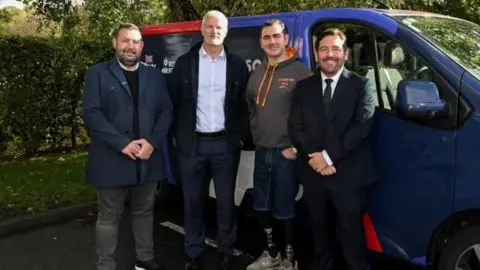 Bernard Platt Four men smile at the camera while standing next to a blue van. The man second from the right is wearing a pair of denim shorts over two prosthetic limbs.