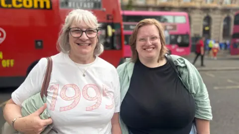 Lillian Lawrence (left) has shoulder length white hair and is wearing a white t-shirt with '1995' printed on it and Katelyn Moore (right) has shoulder length ginger hair and is dressed in a black t-shirt and green shirt. They are pictured on a city centre street-The background is blurred but there are two buses visible. 