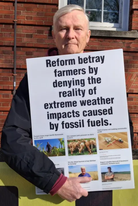 LDRS A man with short grey hair is holding up a sign that says 'Reform betray farmers by denying the reality of extreme weather impacts caused by fossil fuels'. Below the sign are pictures of several BBC articles.