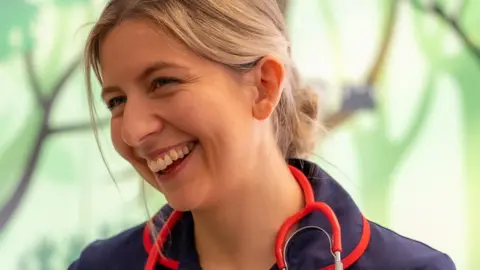 Roald Dahl's Marvellous Children's Charity A woman with blonde hair that is tied back into a bun smiles away from the camera. She is wearing a navy nurse uniform with a red stethoscope around her neck. 