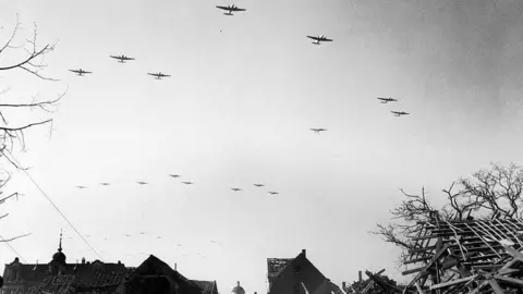 Getty Images Allied planes fly over a shattered German town on their way to drop paratroopers over the Rhine in March 1945. The black and white images shows planes in the sky. Below are roofs with their struts showing, and on the right is a heap of roof struts exposed to the sky. On both sides are leafless tree branches.