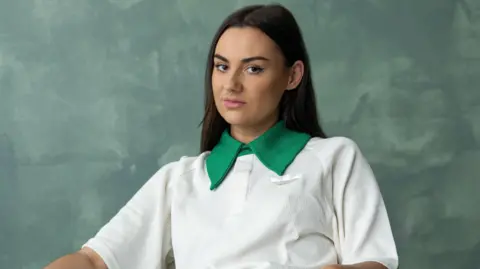Yellowbelly Jade wearing a white and green top stares seriously at the camera