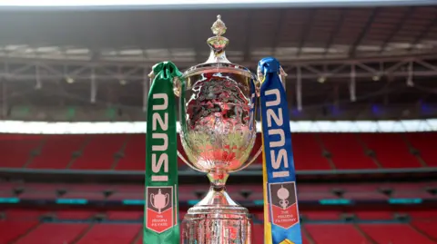 Getty Images The silver trophy of the FA Vase displayed at Wembley Stadium, with empty red seats behind it and blue and green ribbons tied to each handle.