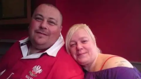 David Taylor A man and a woman are posing together for a photograph. They look happy. He is wearing a Wales rugby shirt and she is wearing a purple off-the-shoulder top.