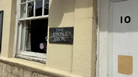A yellow clad building with a white, scuffed door and peeling wooden window frame. A hand-drawn black slate sign reads in white lettering: The Locals Local.