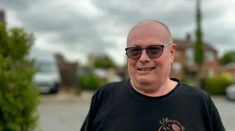 Martin Giles/BBC Christopher Campion is looking at the camera. He is wearing a black T-shirt and spectacles. In the blurred background are houses and a driveway.
