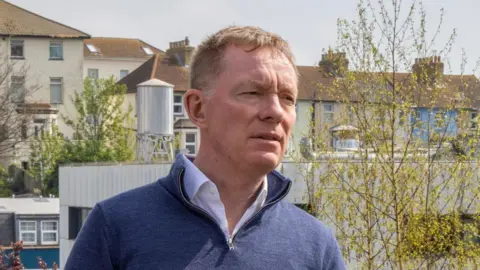 Andrew Aitchinson | Getty Images MP Chris Bryant wears a blue quarter-zip and white shirt as he stands in front of a row of several houses and trees. It is a head and shoulders shot of him and he is looking to the right of the image.