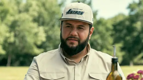 From Dedi Chen/Netflix A Still the Netflix show Mo, showing the main character Mo wearing a cream -colored shirt and hat label "Houston Astros"As he is in front of him in a distance with the top of an olive oil bottle.