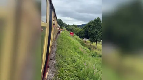 Torquil Buist A shot taken from the window of a vintage train. Passengers can be seen hanging out of the window. People in high-vis jackets stand on the tracks. A red lorry can be seen down an embankment.