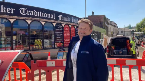 A man in a blue jacket and white T shirt stands in front of orange road closure barriers. He is smiling at the camera. Vans can be seen on the road behind him as well as a shop in the background.