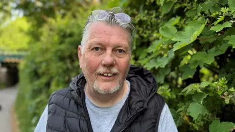 A man wearing a t-shirt and a puffer vest stands smiling in front of a green leafy bush. 