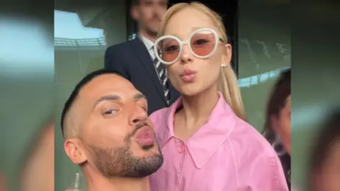 Carl Bembridge poses with the singer Ariana Grande. Carl and Ariana are both pouting and looking at the camera for a selfie. Ariana wears a pink jacket and white rimmed sunglasses.