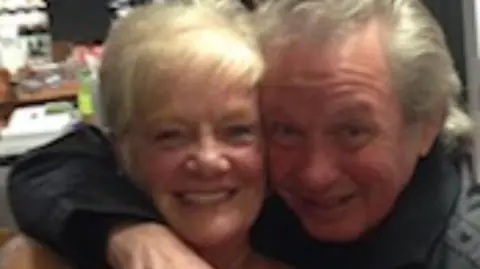 Police handout Sheila Jackson with blond hair smiles as Eric Greener with grey hair has his arm around her neck in front of a bar. Eric is also smiling.