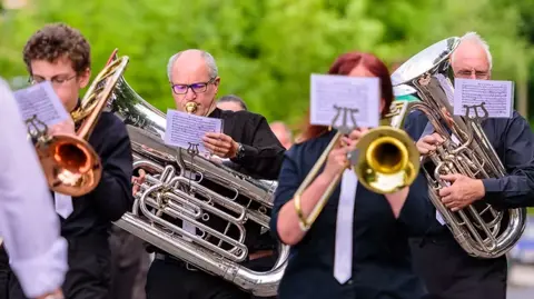 Tameside Whit Friday Band Contest A number of people in black shirts and white ties play brass instruments while walking outside. There is sheet music attached to each person's instrument.