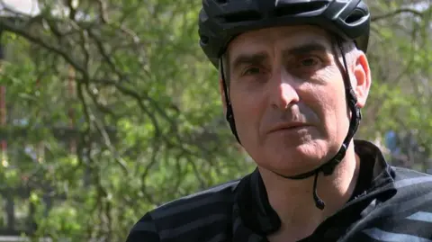 A man in a dark top wearing a cycling helmet is looking at the camera. Leaves and branches are blurred in the background.