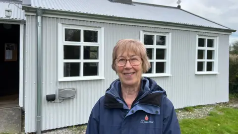 Maureen Phillips is standing outside the little tin church. She is wearing a blue jacket and has short hair. The church has a tin exterior and three windows on the side.